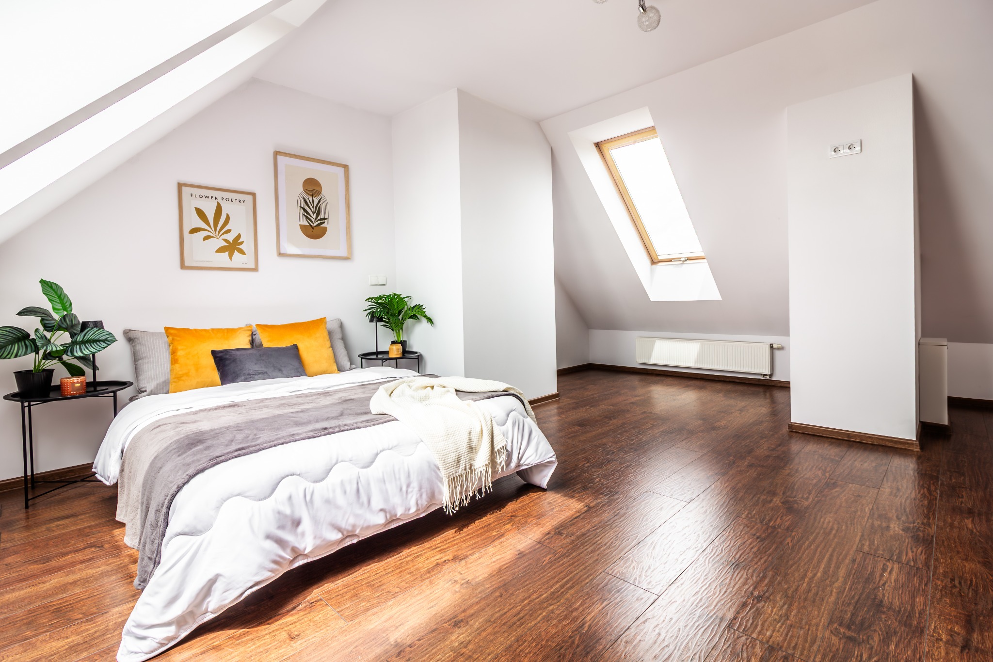 Jasne poddasze z drewnianą podłogą i oknem dachowym. Ustawione łóżko z roślinami po bokach. Minimalistyczne plakaty na ścianie dodają uroku.