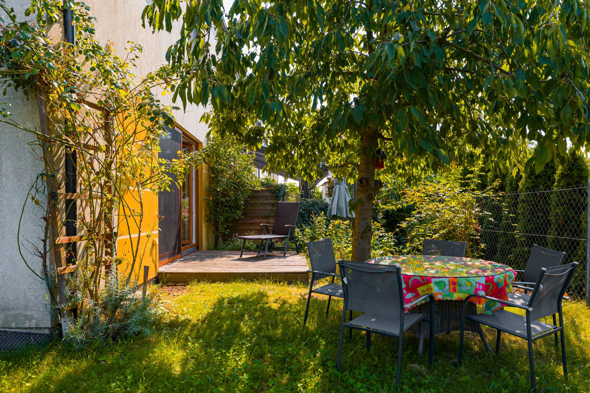 Urokliwy ogród z żółtą elewacją domu, drewnianym tarasem i okrągłym stołem pod rozłożystym drzewem. Krzesła otaczają stół nakryty kolorowym obrusem.