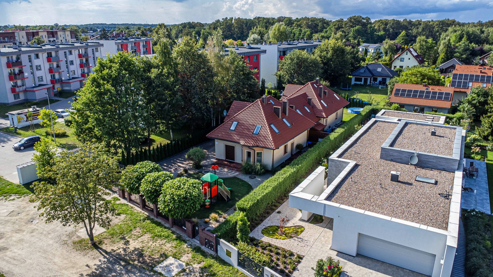 Panoramiczny widok z góry na osiedle domów jednorodzinnych z czerwonymi dachami, zielenią i placem zabaw. W tle bloki mieszkalne i las.