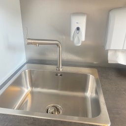 Viktoriia Polishchuk - Stalowa umywalka z baterią, dozownik do mydła marki Faneco i podajnik ręczników papierowych na tle metalowej ściany w pomieszczeniu.