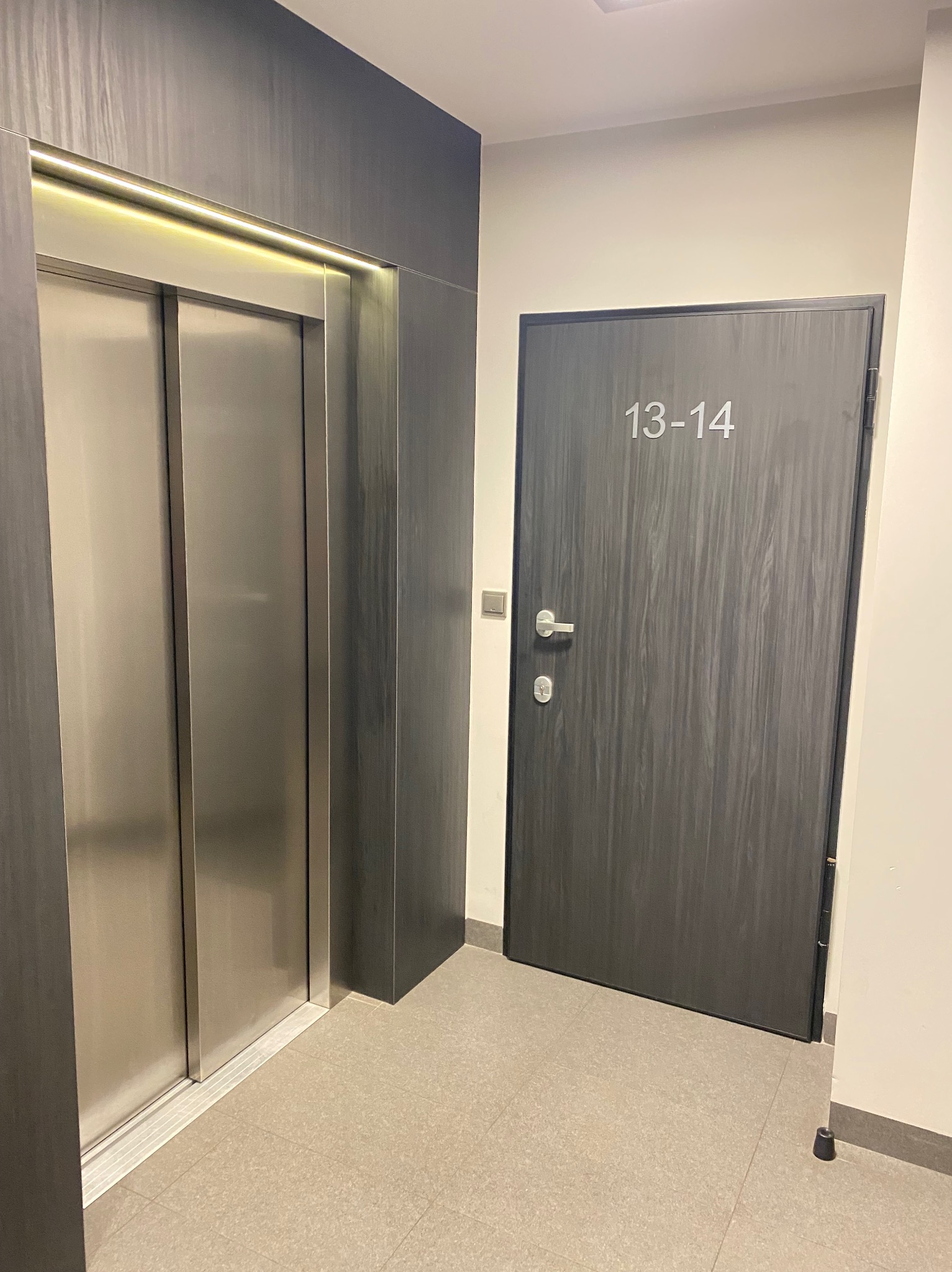 Wnętrze korytarza z drzwiami o numerze 13-14 i windą z podświetleniem LED, utrzymane w stonowanej kolorystyce szarości i beżu.