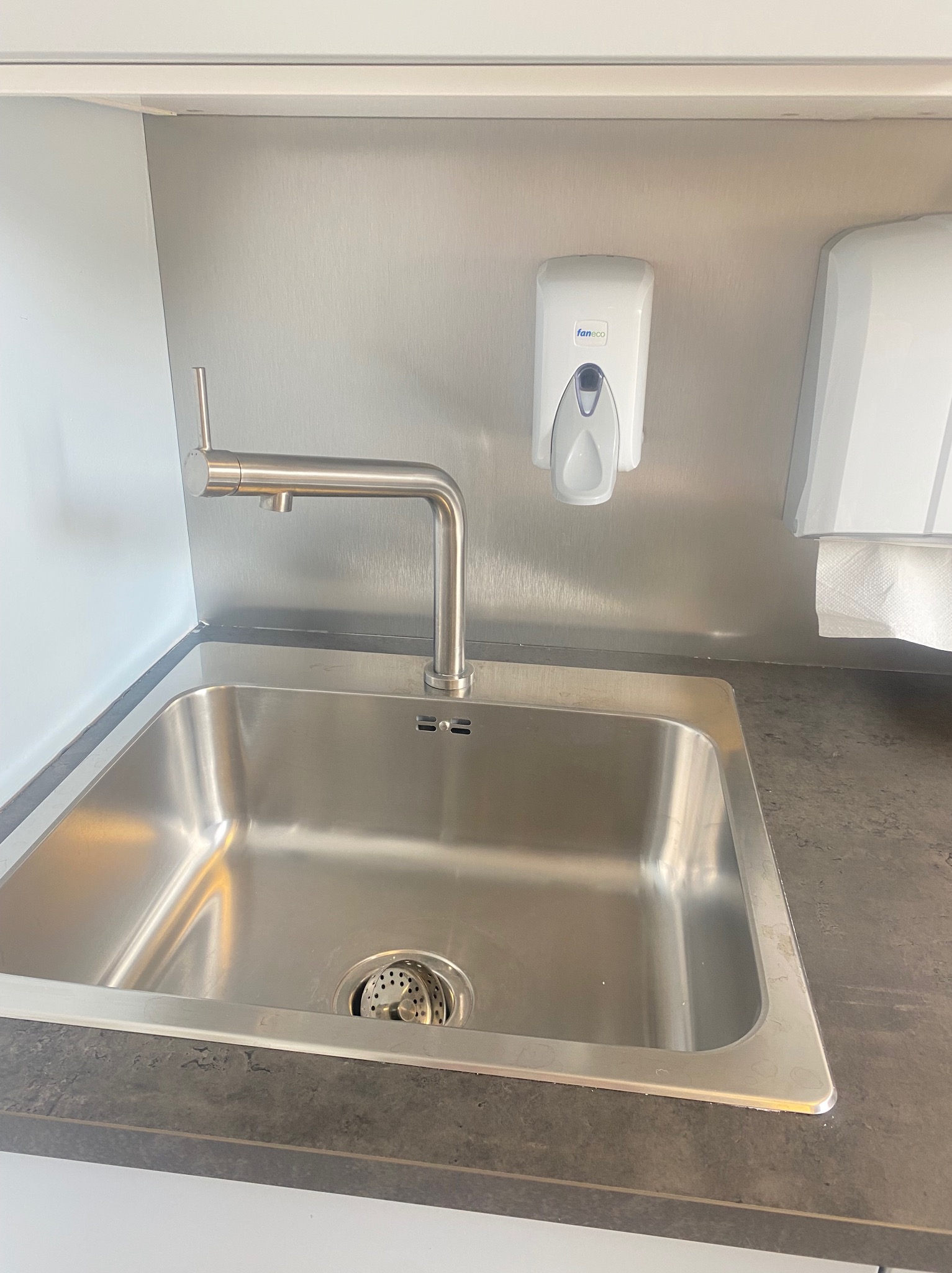 Stalowa umywalka z baterią, dozownik do mydła marki Faneco i podajnik ręczników papierowych na tle metalowej ściany w pomieszczeniu.