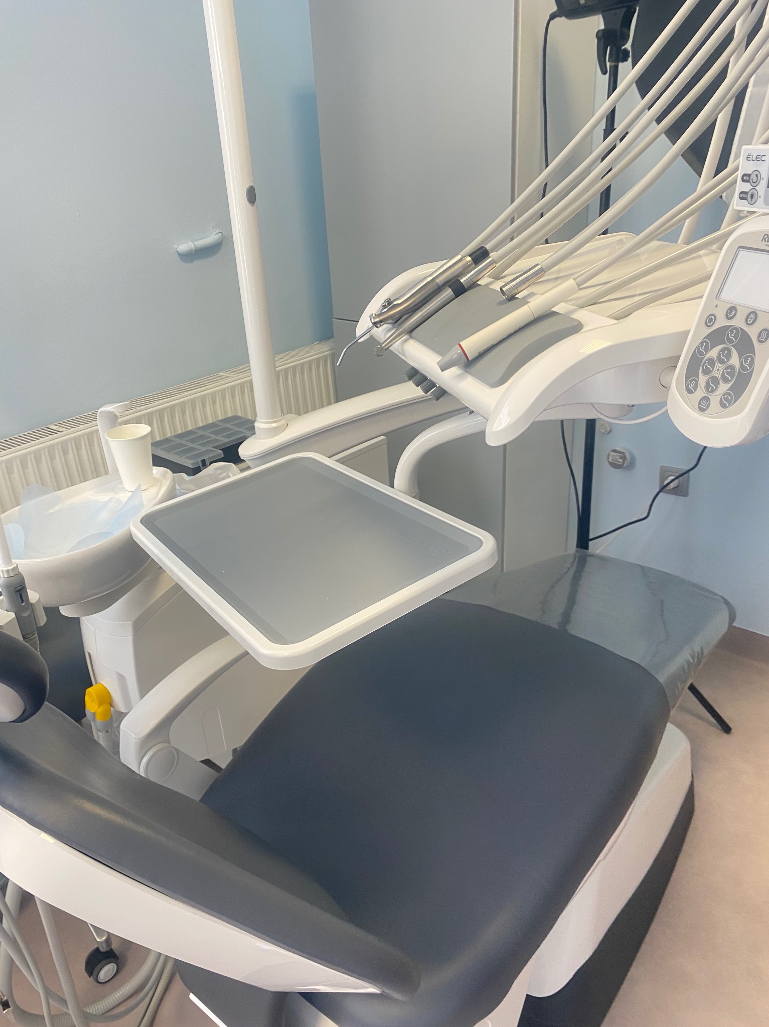 Nowoczesne, biało-szare stanowisko dentystyczne z licznymi przewodami i panelem sterowania, gotowe do przyjęcia pacjenta, z kubkiem i podkładem na tacy.