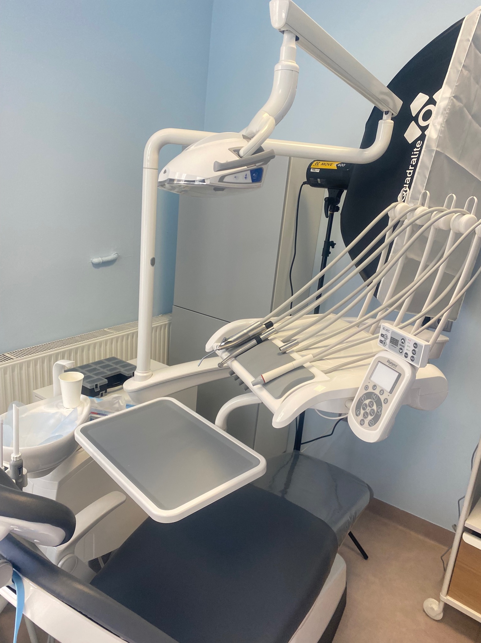 Białe, nowoczesne krzesło dentystyczne z licznymi wężami i panelem sterowania, ustawione w jasnym gabinecie z niebieskimi ścianami i lampą oświetleniową w tle.