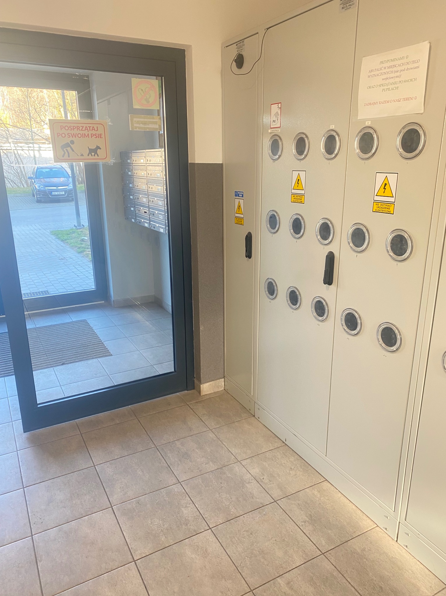 Wejście do budynku z tabliczką przypominającą o sprzątaniu po psie, widoczne skrzynki pocztowe i szafa elektryczna z ostrzeżeniami.