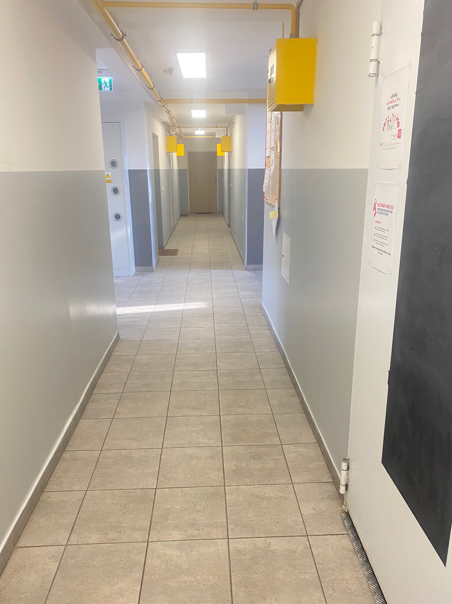 Długi, pusty korytarz w budynku z żółtymi rurami pod sufitem i plakatami na ścianach, widziany z perspektywy osoby wchodzącej.