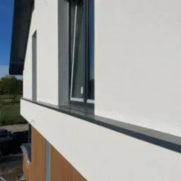 Otwarty, grafitowy profil okienny w nowym budynku z białą elewacją w Warce. Widoczne elementy wykończeniowe i przygotowania do dalszych prac.