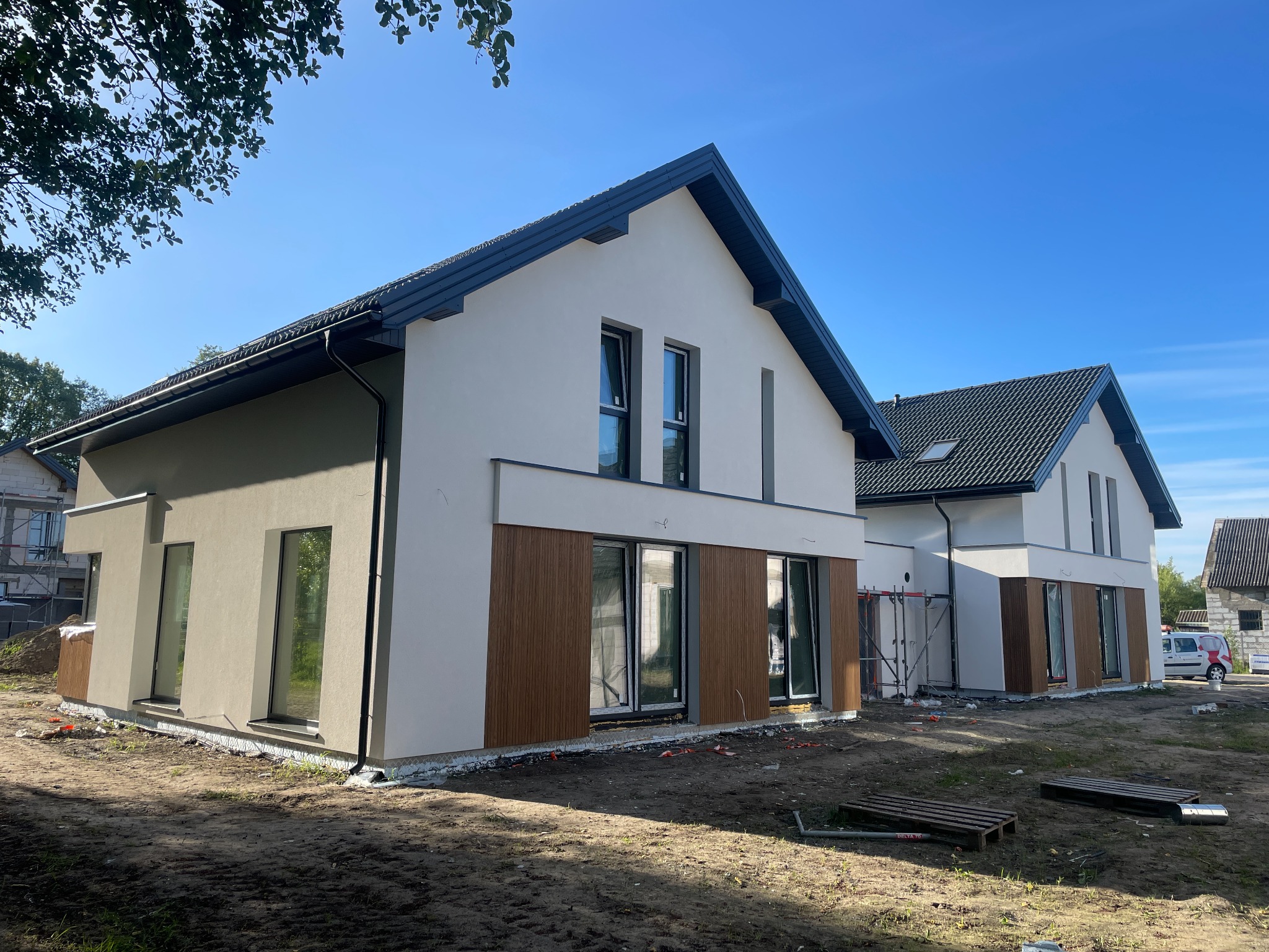 Nowoczesny, dwukondygnacyjny dom jednorodzinny z oknami i drewnianą elewacją, w trakcie wykończenia. Widoczne rusztowania i teren budowy.