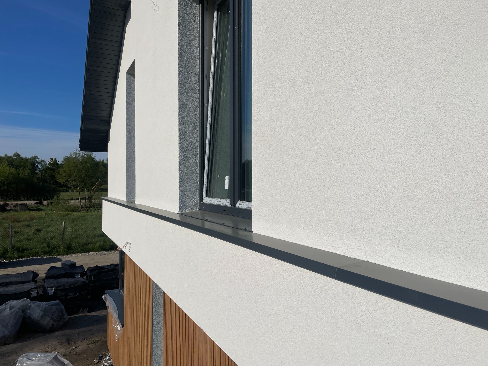 Otwarty, grafitowy profil okienny w nowym budynku z białą elewacją w Warce. Widoczne elementy wykończeniowe i przygotowania do dalszych prac.
