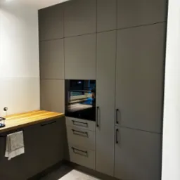 Nowoczesna kuchnia w odcieniach szarości z drewnianym blatem i czarnymi uchwytami, wbudowany piekarnik. Minimalistyczny design z oświetleniem punktowym.