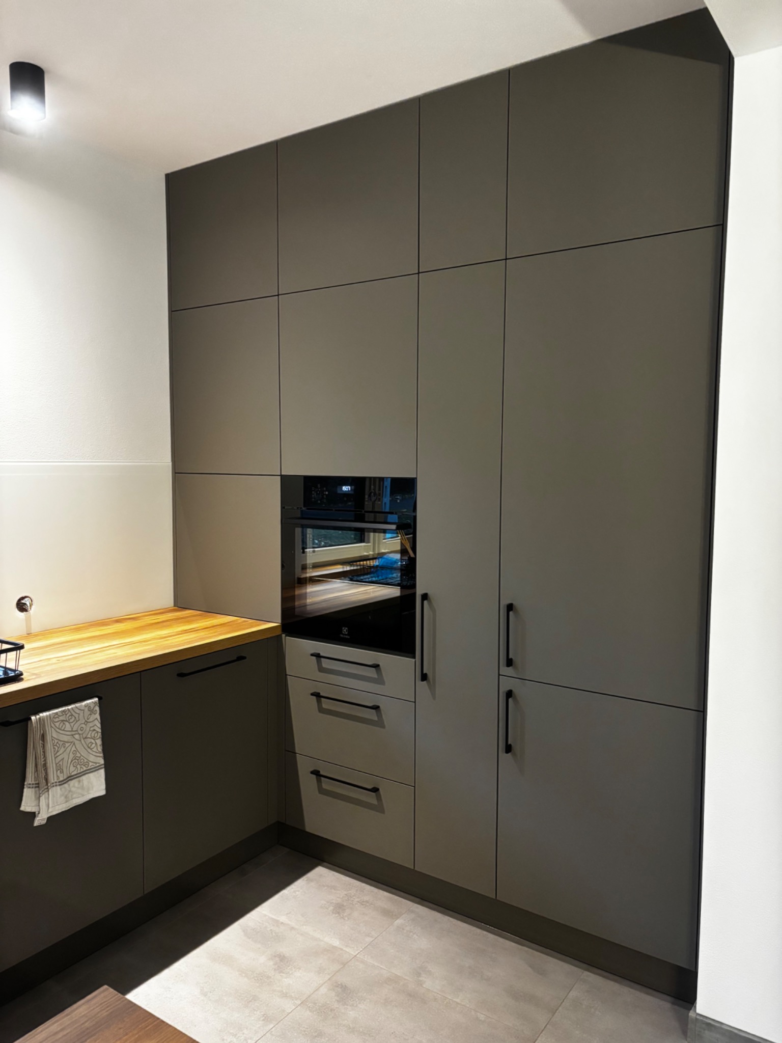 Nowoczesna kuchnia w odcieniach szarości z drewnianym blatem i czarnymi uchwytami, wbudowany piekarnik. Minimalistyczny design z oświetleniem punktowym.