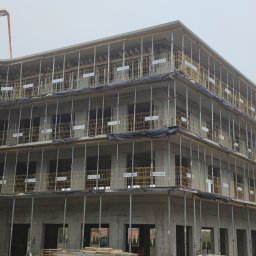 Triumh - Wielopiętrowy budynek żelbetowy w trakcie budowy, widoczne szalunki i zbrojenia, dźwig budowlany w tle. Ujęcie z dołu, pochmurna pogoda.