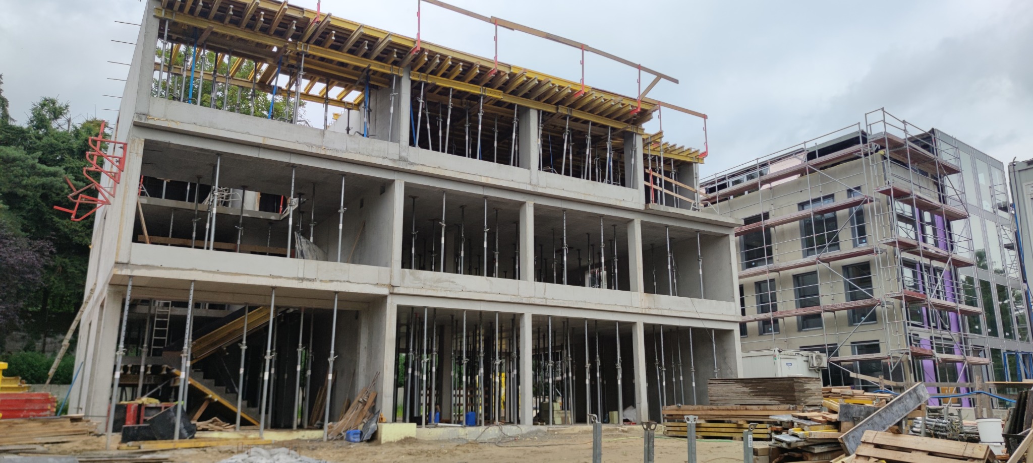 Surowy stan budynku żelbetowego w trakcie budowy, widoczne podpory i szalunki, sąsiedni budynek w rusztowaniach, pochmurne niebo w tle.