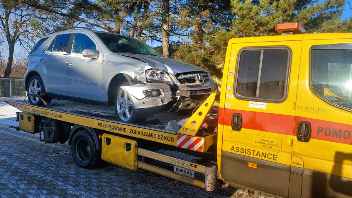 Uszkodzony srebrny Mercedes ML na żółtej lawecie z napisem 'Centrum Likwidacji Szkód Poznań' na tle zaśnieżonego terenu i drzew.
