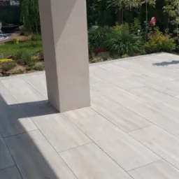 Jasne, prostokątne płyty tarasowe ułożone na zewnątrz, z widocznym słupem konstrukcyjnym i fragmentem ścieżki z kostki. W tle zieleń i elementy małej architektury.