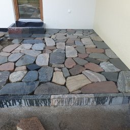 Usługi Brukarskie Ogólno budowlane - Układanie nieregularnej kostki granitowej w odcieniach szarości i brązu przy ścianie budynku. Widoczne elementy wózka budowlanego i kamienne obrzeże.