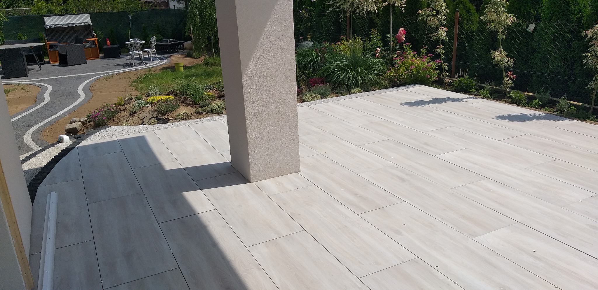 Jasne, prostokątne płyty tarasowe ułożone na zewnątrz, z widocznym słupem konstrukcyjnym i fragmentem ścieżki z kostki. W tle zieleń i elementy małej architektury.