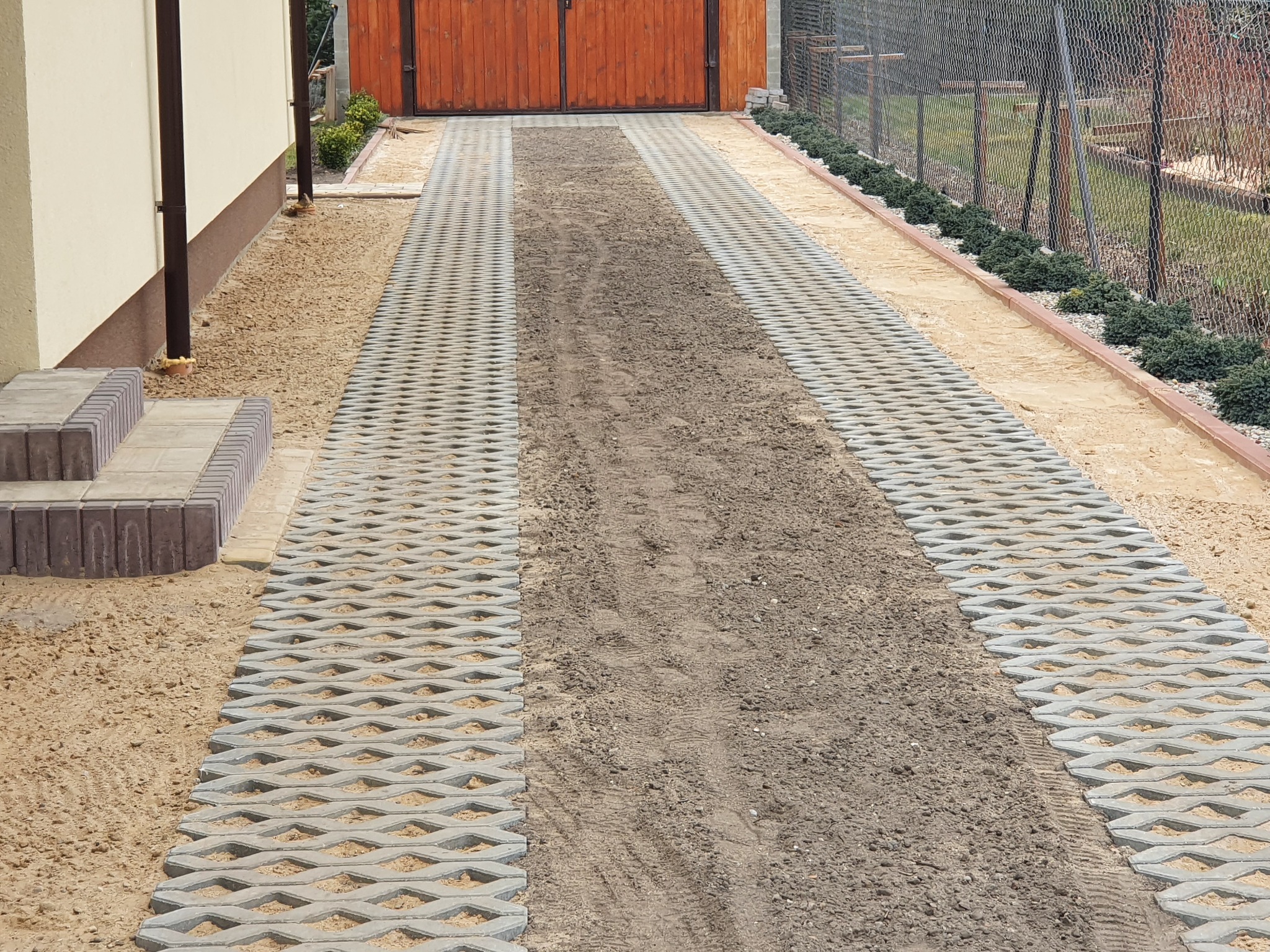 Ułożona ażurowa kostka brukowa na podjeździe, z częściowo wypełnionymi przestrzeniami. Widoczne schody z kostki i ogrodzenie z siatki. Podłoże z piasku.