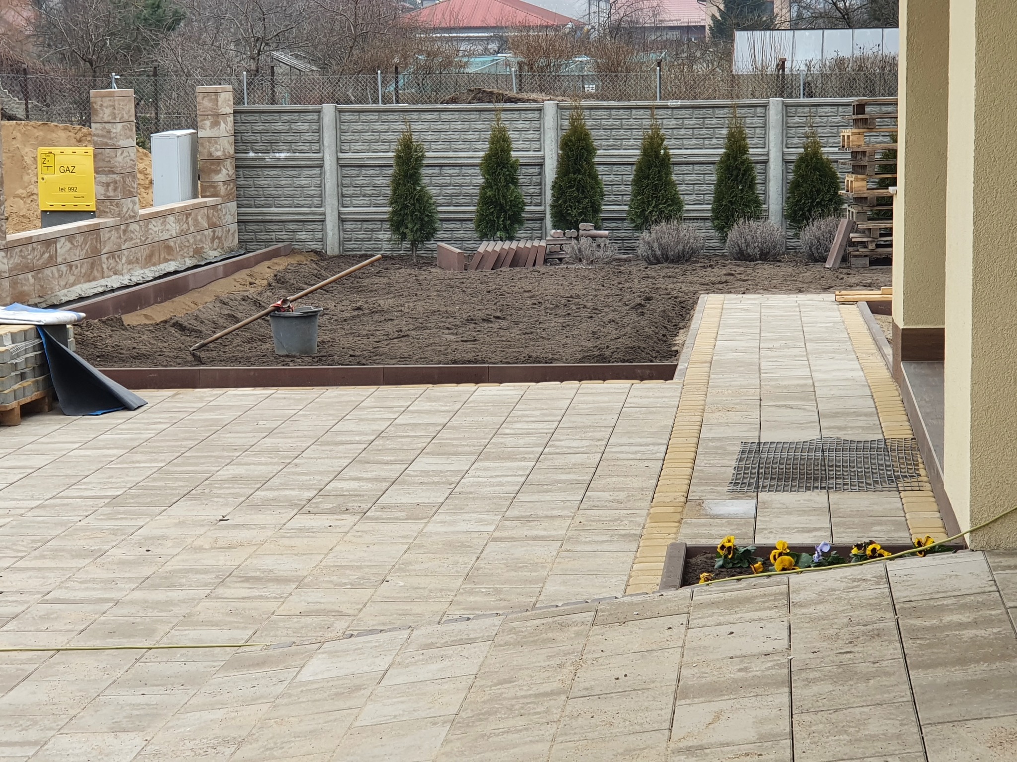 Wykończony taras z kostki brukowej w ogrodzie, z fragmentem rabaty z tujami i kostką do dalszego układania. Widoczne elementy ogrodzenia i skrzynka gazowa.