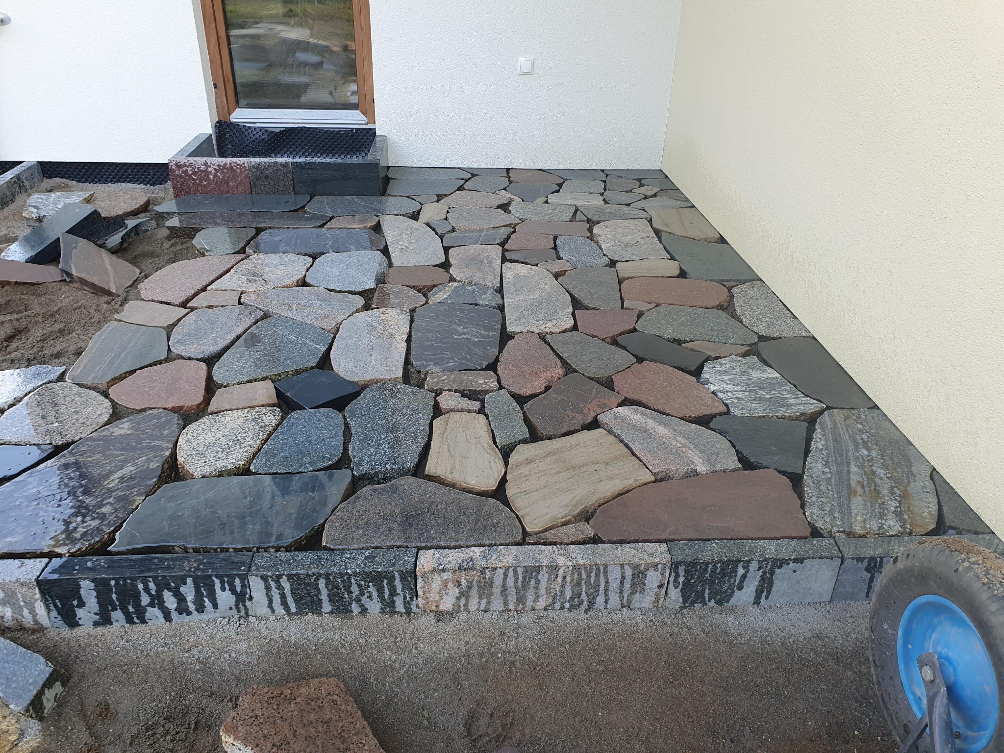 Układanie nieregularnej kostki granitowej w odcieniach szarości i brązu przy ścianie budynku. Widoczne elementy wózka budowlanego i kamienne obrzeże.
