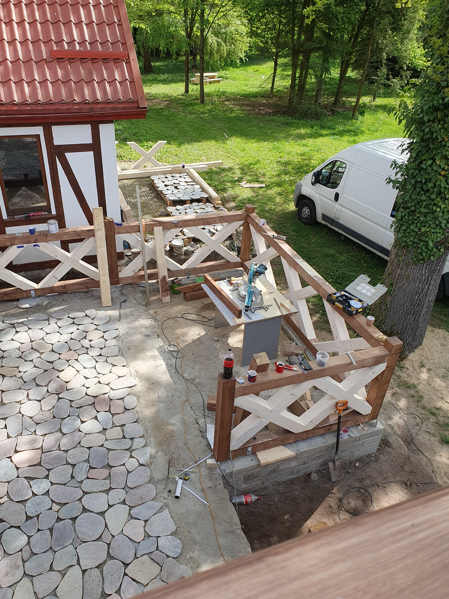 Budowa drewnianej balustrady z dekoracyjnym wzorem krzyżowym i układanie ścieżki z nieregularnych kamieni na posesji z budynkiem i samochodem dostawczym w tle.