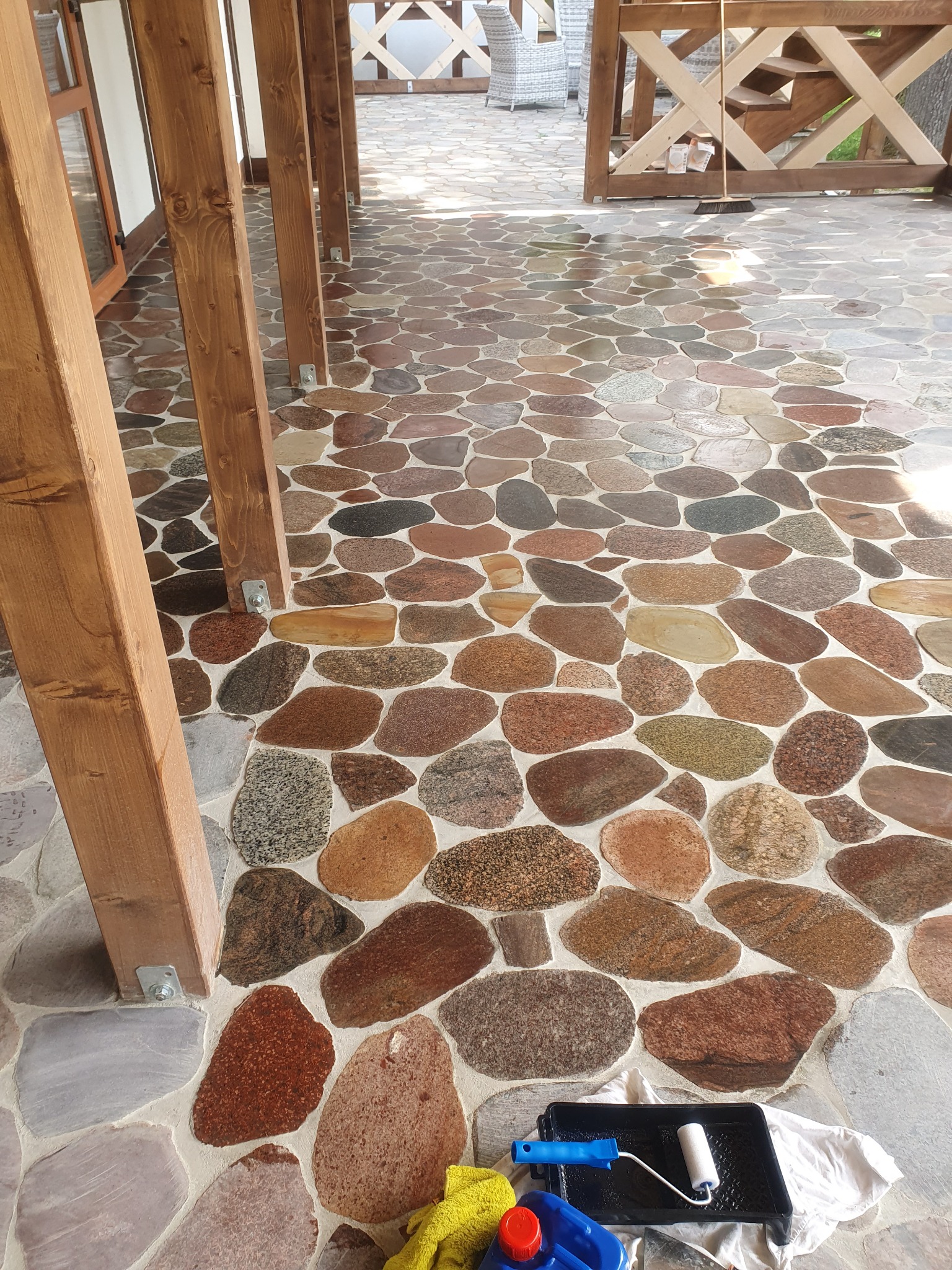 Balkon z podłogą z nieregularnych kamieni w odcieniach brązu i szarości. Widoczne drewniane słupy, narzędzia malarskie i fragment balustrady w tle.