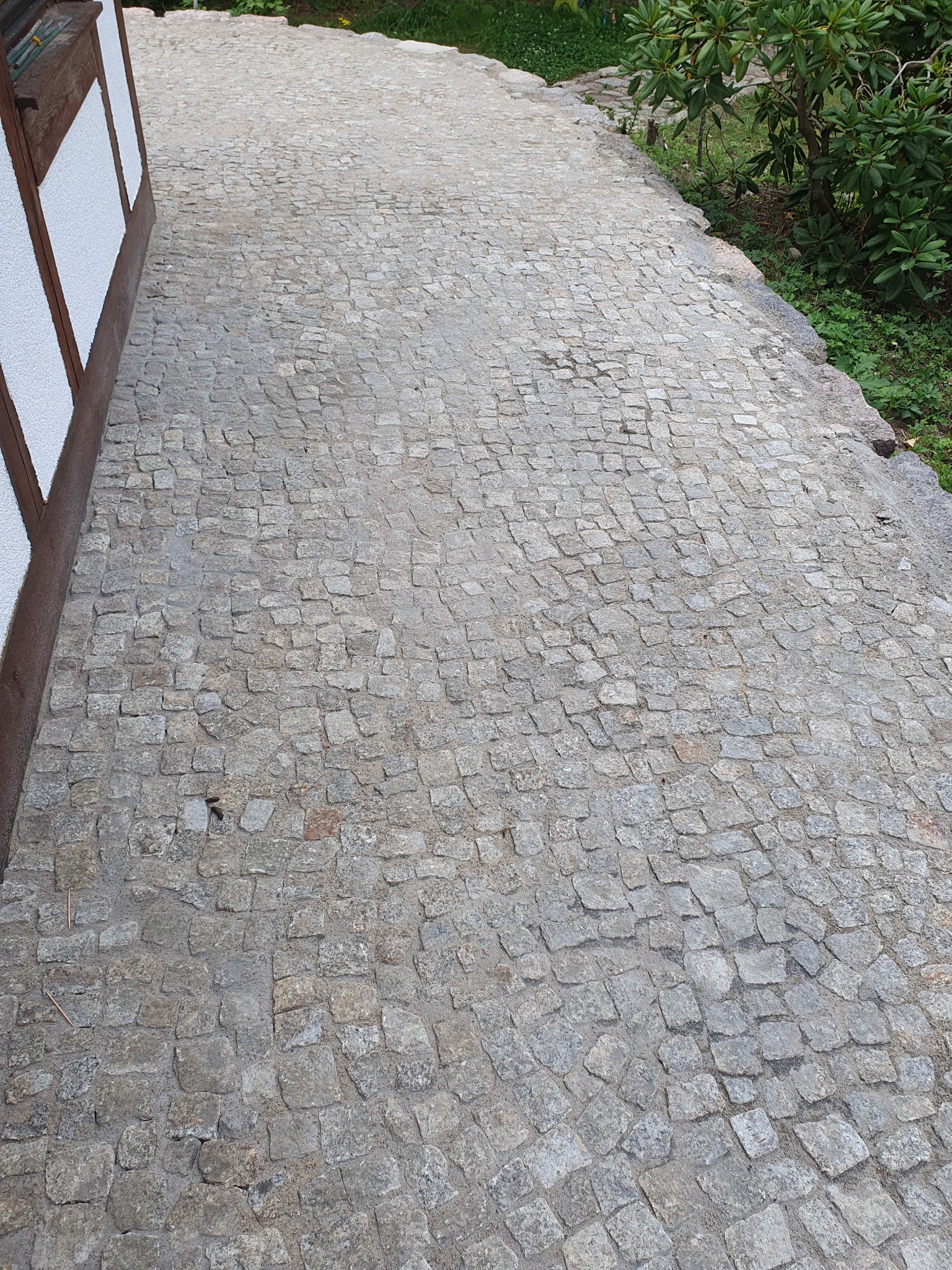 Ścieżka z szarej kostki granitowej ułożona przy budynku z elementami drewnianymi i zielenią. Widok z góry, ukazujący detale faktury kamienia i precyzję wykonania.