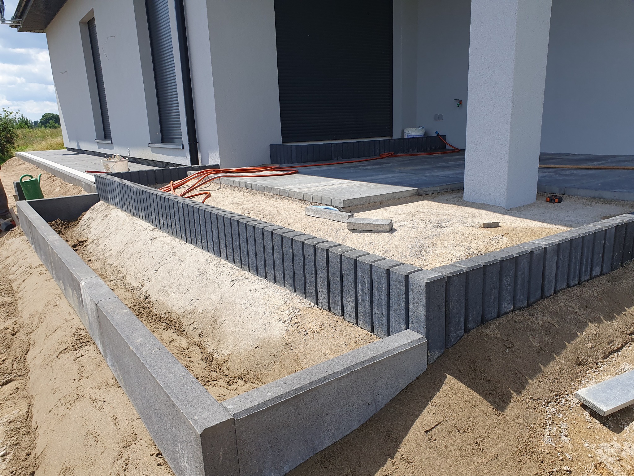 Etap prac: obrzeża z kostki i częściowo ułożona kostka brukowa na posesji z nowoczesnym domem. Widoczne przewody i piasek przygotowany pod dalsze prace.