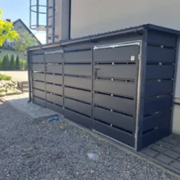 Nowoczesna, antracytowa zabudowa śmietnikowa z poziomych paneli, przylegająca do budynku. Widoczne kamienie, kostka brukowa i fragment zieleni.