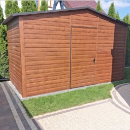 Firma Produkcyjno-Usługowo-Handlowa KamBlach Kamil Trzupek - Brązowy garaż blaszany z imitacją drewna, zamykany na klucz, na tle zieleni i kostki brukowej. Na dachu widoczna osoba.