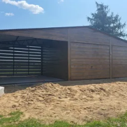 Drewnopodobny, otwarty garaż z metalową konstrukcją w Szczyrzycu. Widoczne elementy konstrukcyjne i boczna ściana z poziomych desek. Teren przygotowany pod wylewkę.