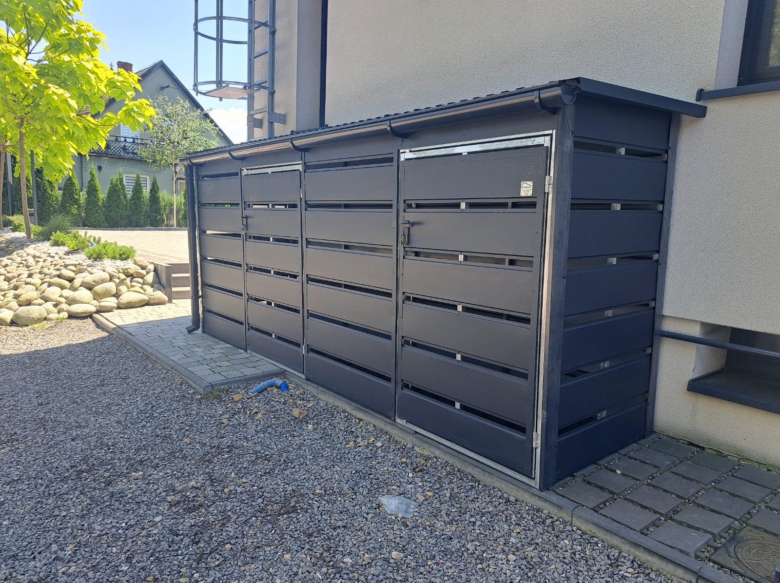 Nowoczesna, antracytowa zabudowa śmietnikowa z poziomych paneli, przylegająca do budynku. Widoczne kamienie, kostka brukowa i fragment zieleni.