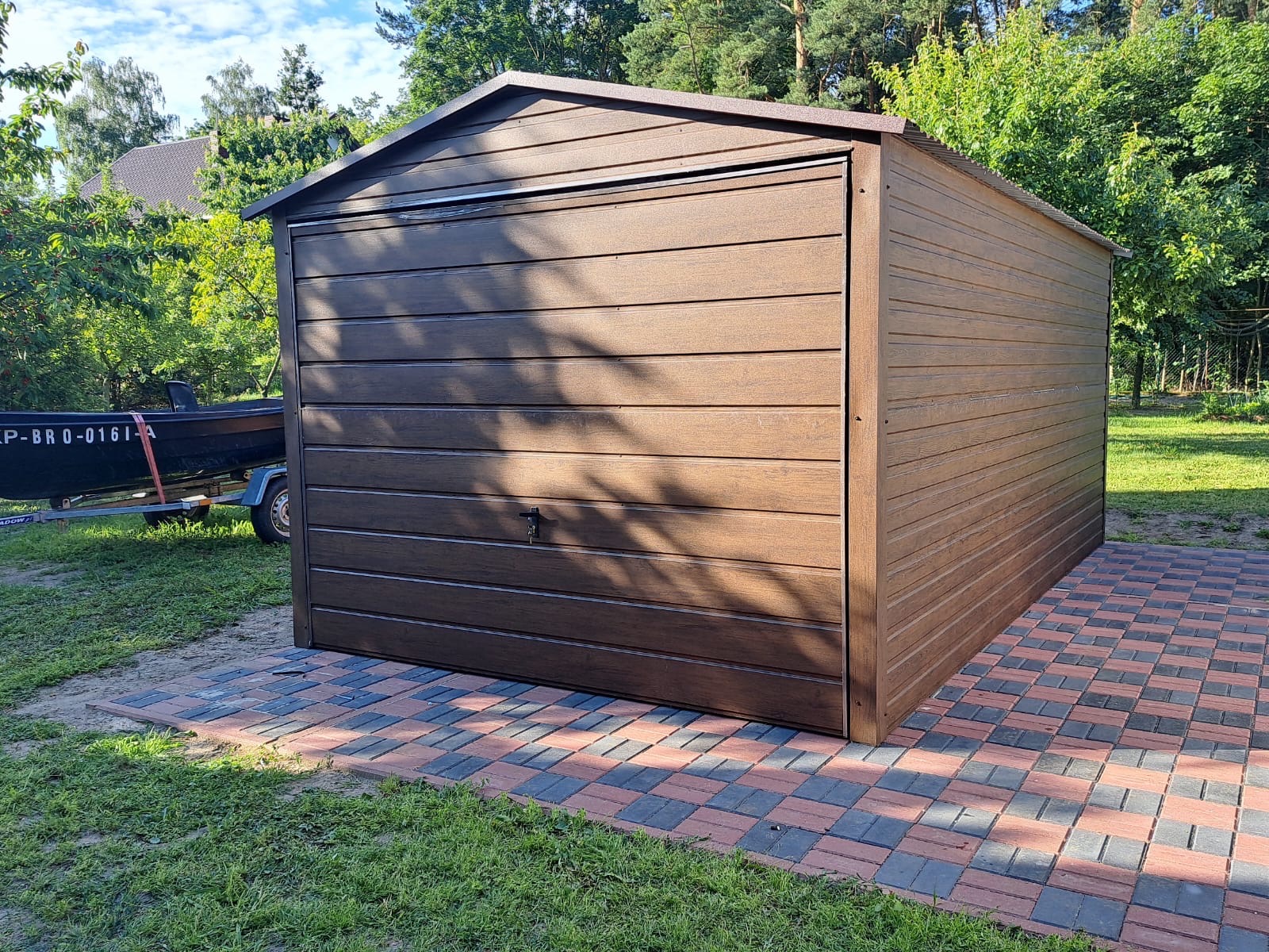 Gotowy garaż blaszany w Szczyrzycu z imitacją drewna, ustawiony na kostce brukowej. W tle zieleń drzew i przyczepa z łodzią.