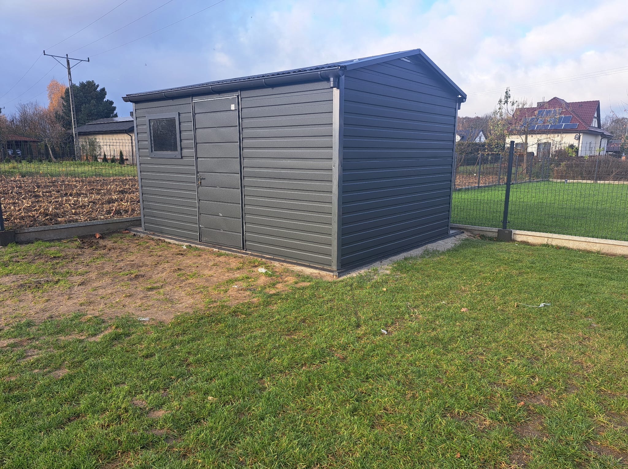 Nowoczesny, grafitowy garaż blaszany z oknem i drzwiami, ustawiony na trawniku w ogrodzie. W tle widoczny dom z panelami słonecznymi i pole uprawne.
