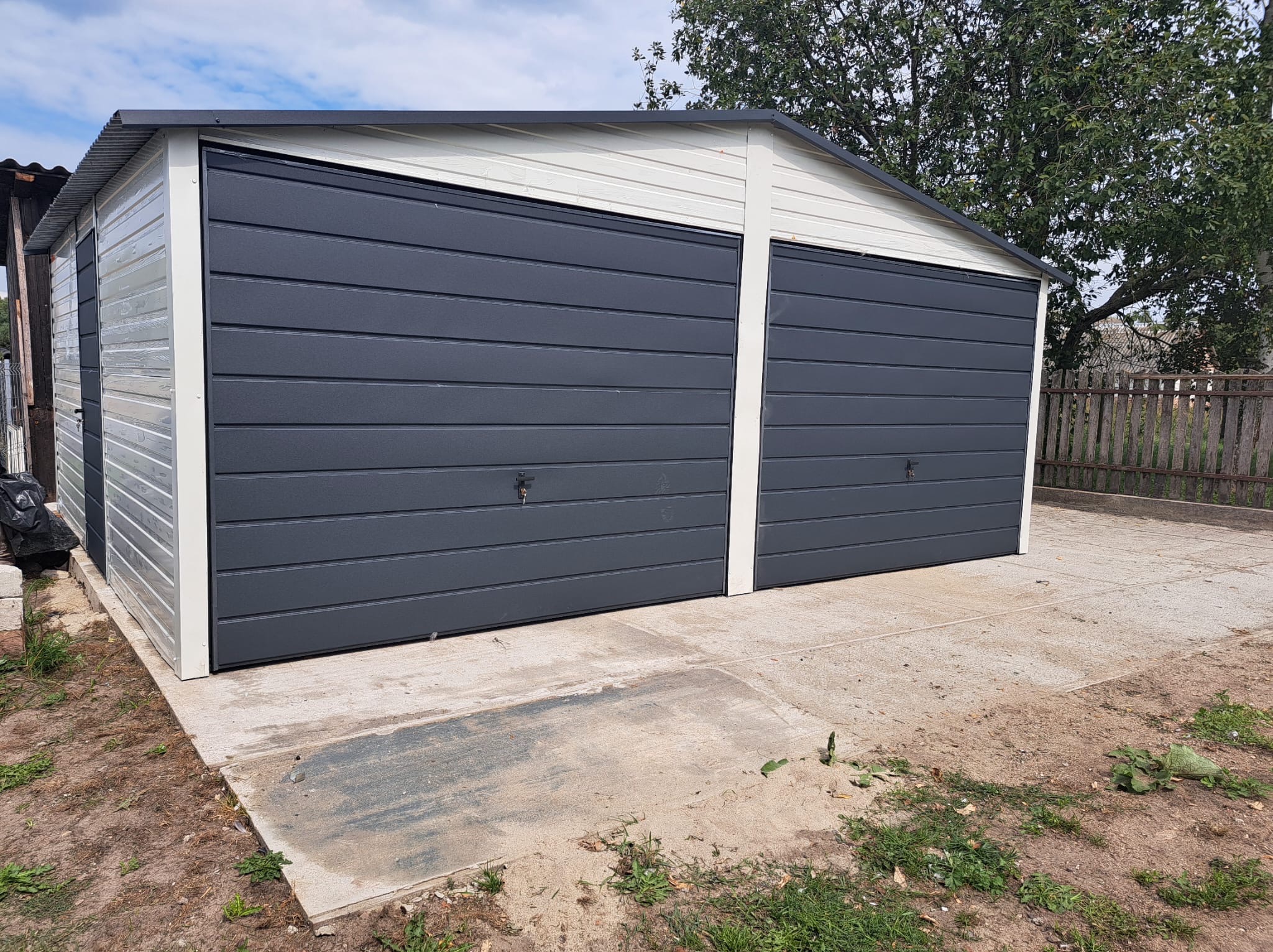 Dwustanowiskowy garaż blaszany z szarymi, segmentowymi bramami, umieszczony na betonowej wylewce. Widok na garaż z zewnątrz, w tle drzewa i płot.