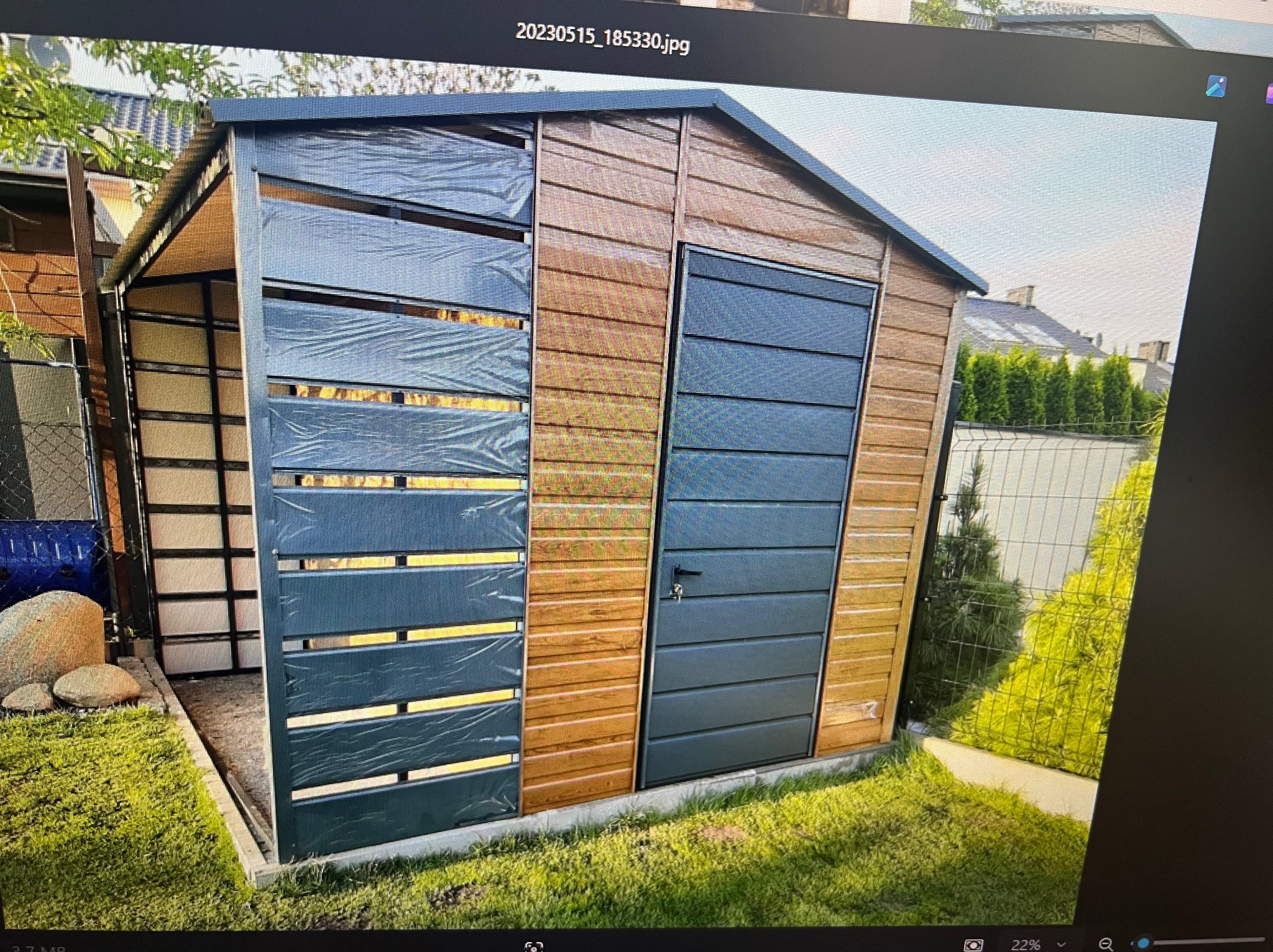 Nowoczesny garaż blaszany z drewnianą elewacją i szarą bramą segmentową, otoczony zielonym trawnikiem i ogrodzeniem z siatki.