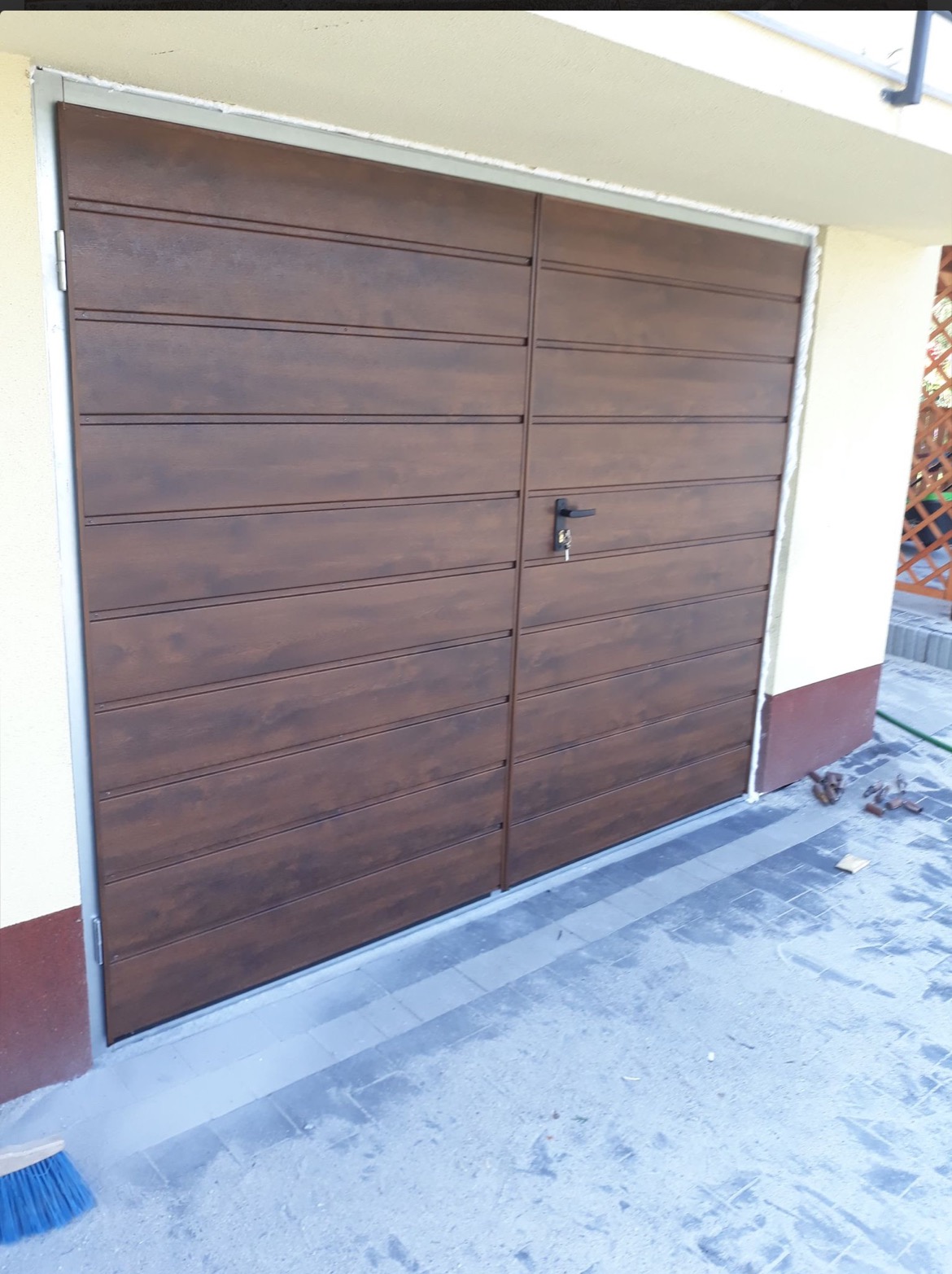 Nowa, brązowa brama garażowa segmentowa z poziomym wzorem paneli, z zamkiem na klucz, zamontowana w jasnym budynku. Widoczny fragment kostki brukowej i szczotka.