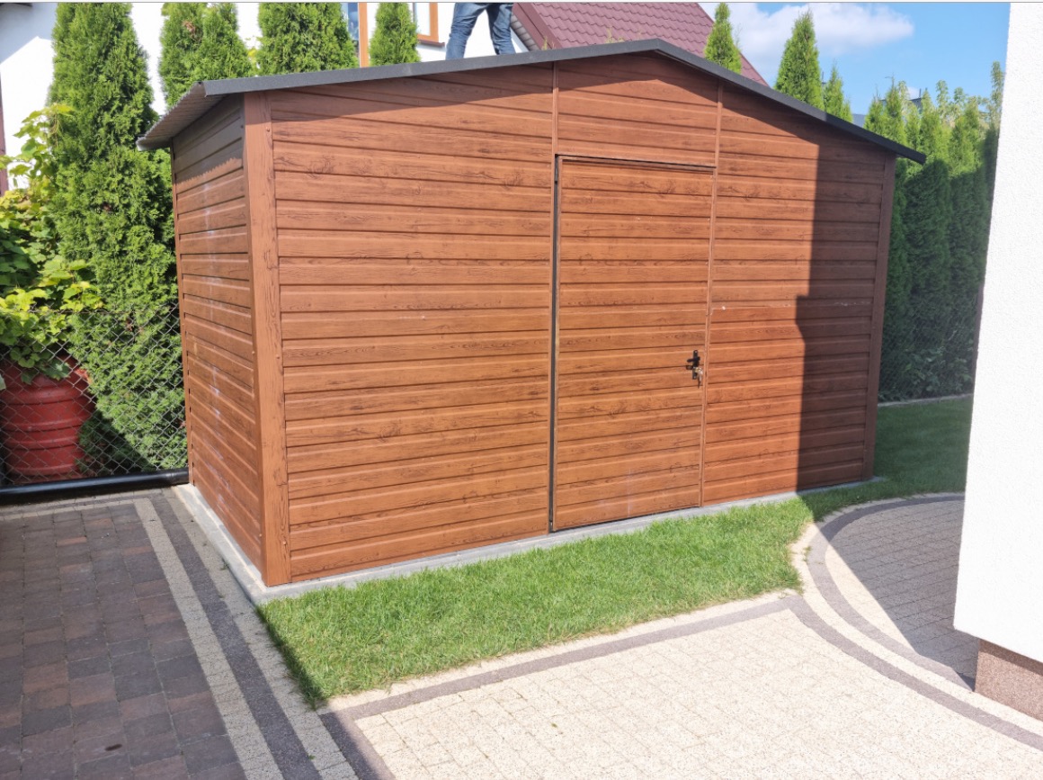Brązowy garaż blaszany z imitacją drewna, zamykany na klucz, na tle zieleni i kostki brukowej. Na dachu widoczna osoba.