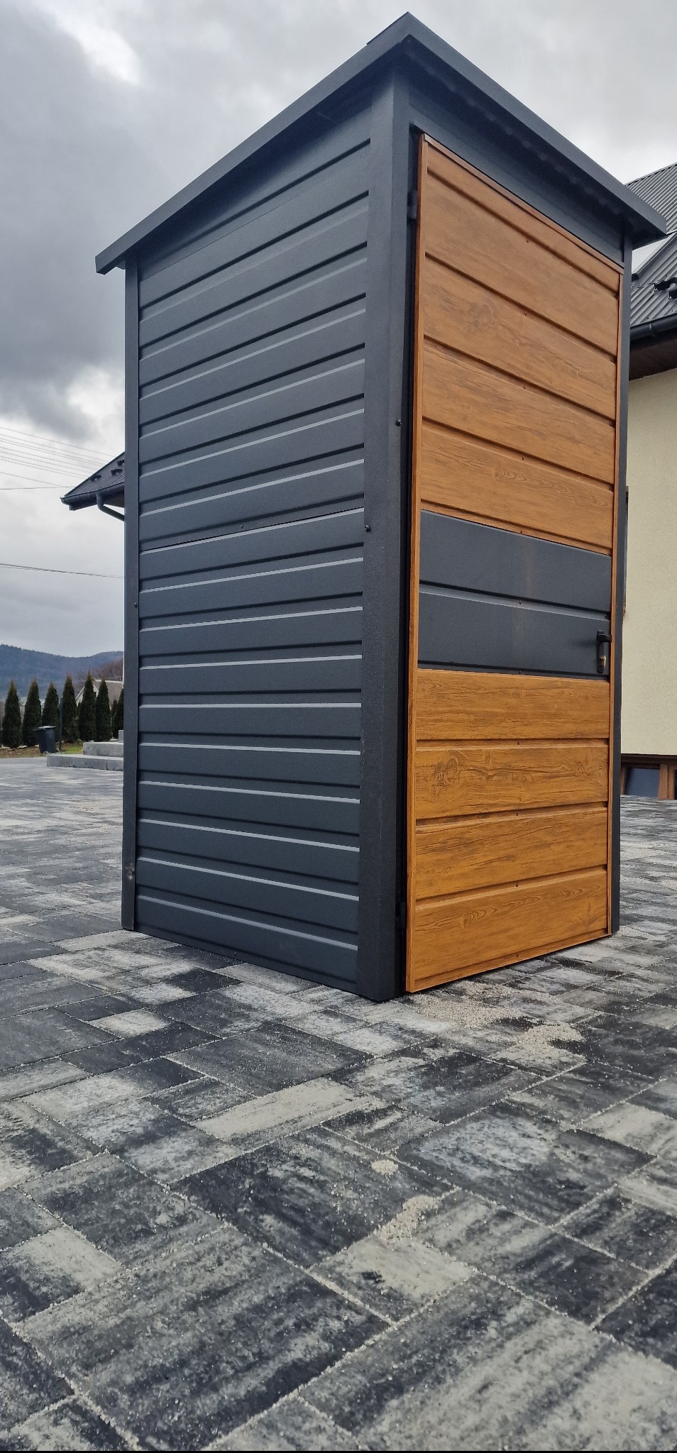 Nowoczesna, metalowa szafa ogrodowa z drewnopodobną, dwukolorową elewacją, stojąca na szarej kostce brukowej. Widoczne góry i zabudowa jednorodzinna w tle.