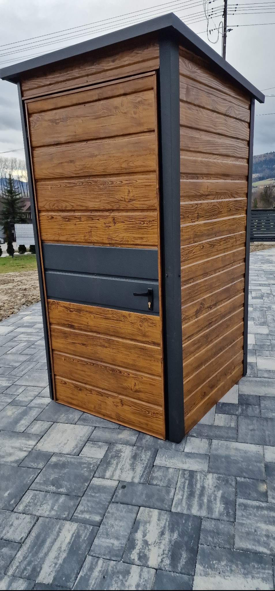 Drewnopodobny, metalowy garaż blaszany z drzwiami i czarną ramą, ustawiony na szarej kostce brukowej. W tle widoczne drzewa i góry.