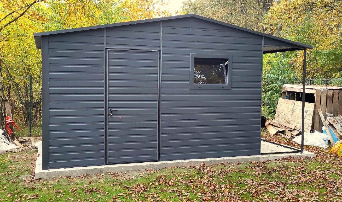 Nowoczesny, antracytowy garaż blaszany z oknem i drzwiami, posadowiony na betonowej wylewce, na tle jesiennego ogrodu.