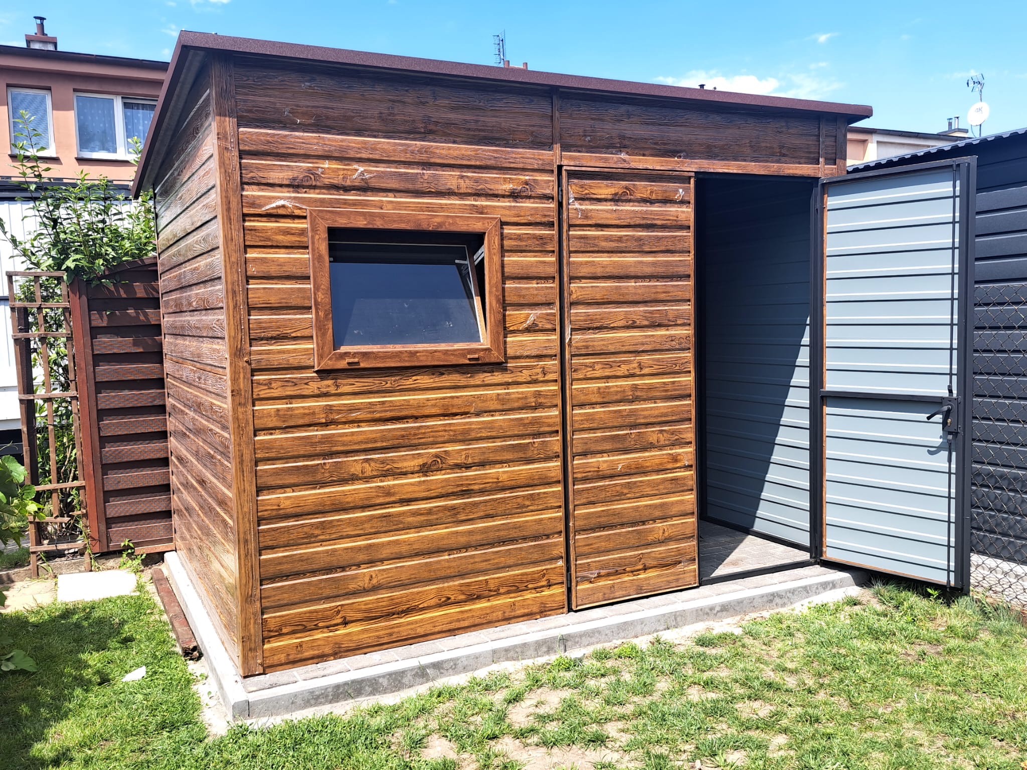 Drewniany domek narzędziowy z oknem i otwartymi drzwiami, umieszczony na betonowej podstawie w ogrodzie. Widoczne deski z wyraźnym rysunkiem słojów.