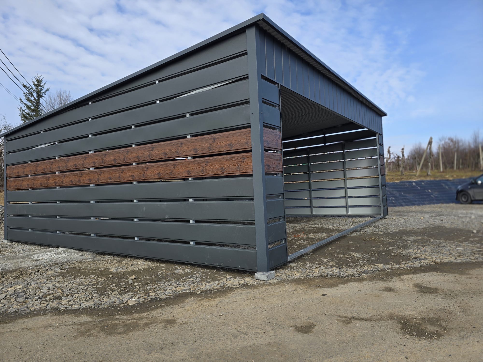Nowoczesny garaż blaszany z elementami dekoracyjnymi imitującymi drewno, na żwirowym podłożu. Widok z boku, w tle błękitne niebo i fragment krajobrazu.