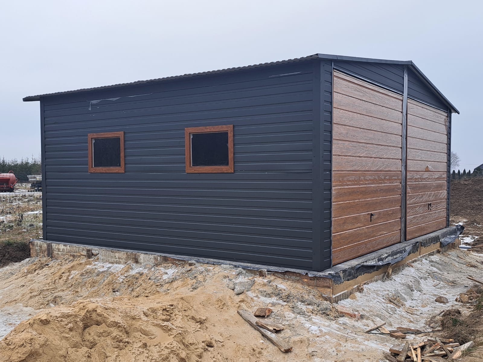 Gotowy garaż blaszany z oknami i bramą imitującą drewno, posadowiony na fundamencie. Widoczne ślady prac ziemnych wokół konstrukcji i elementy drewniane.