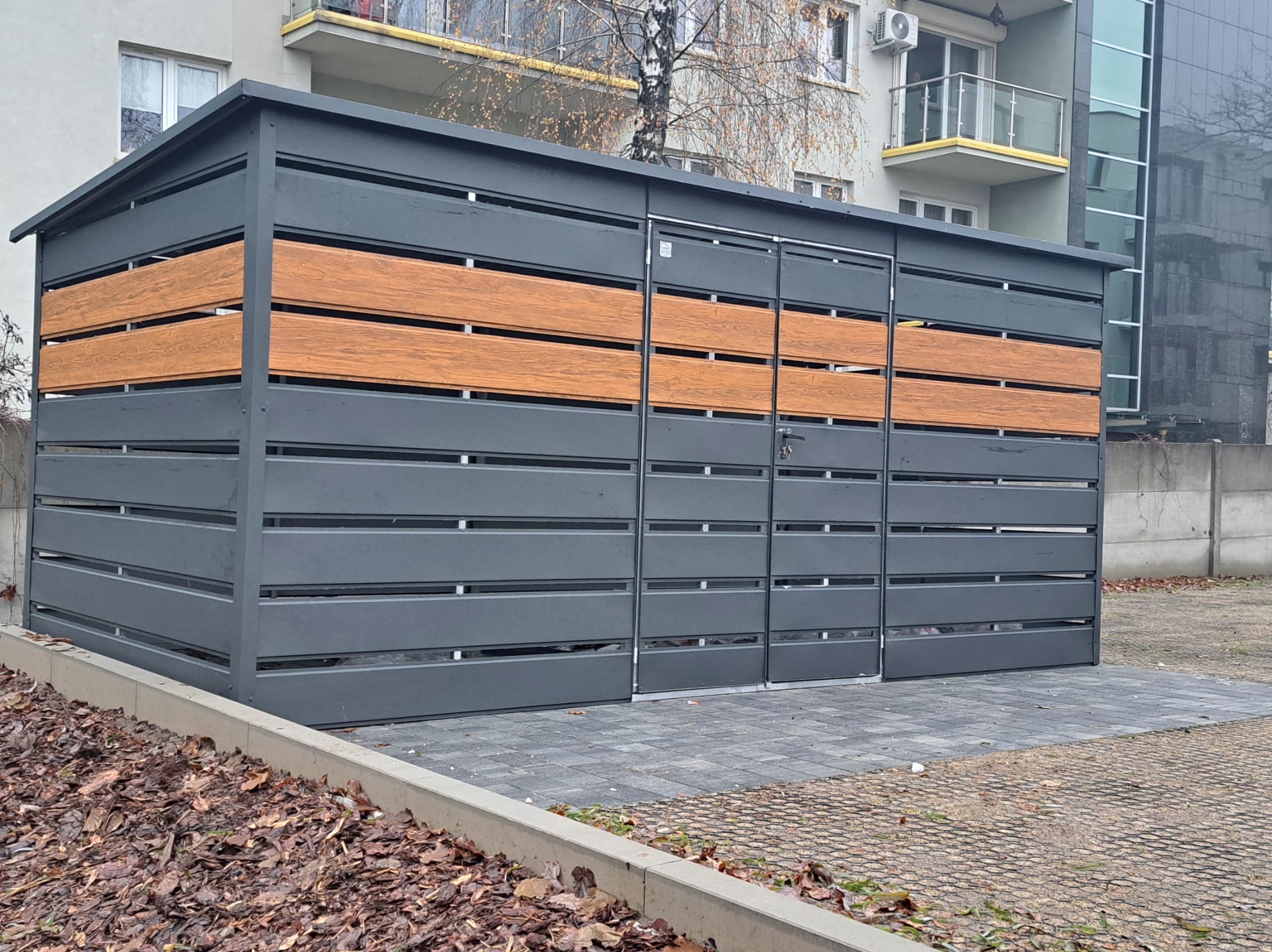 Nowoczesny, grafitowy garaż blaszany z poziomymi, drewnopodobnymi panelami. Dwuspadowy dach, zamek w drzwiach. Na tle bloku mieszkalnego.