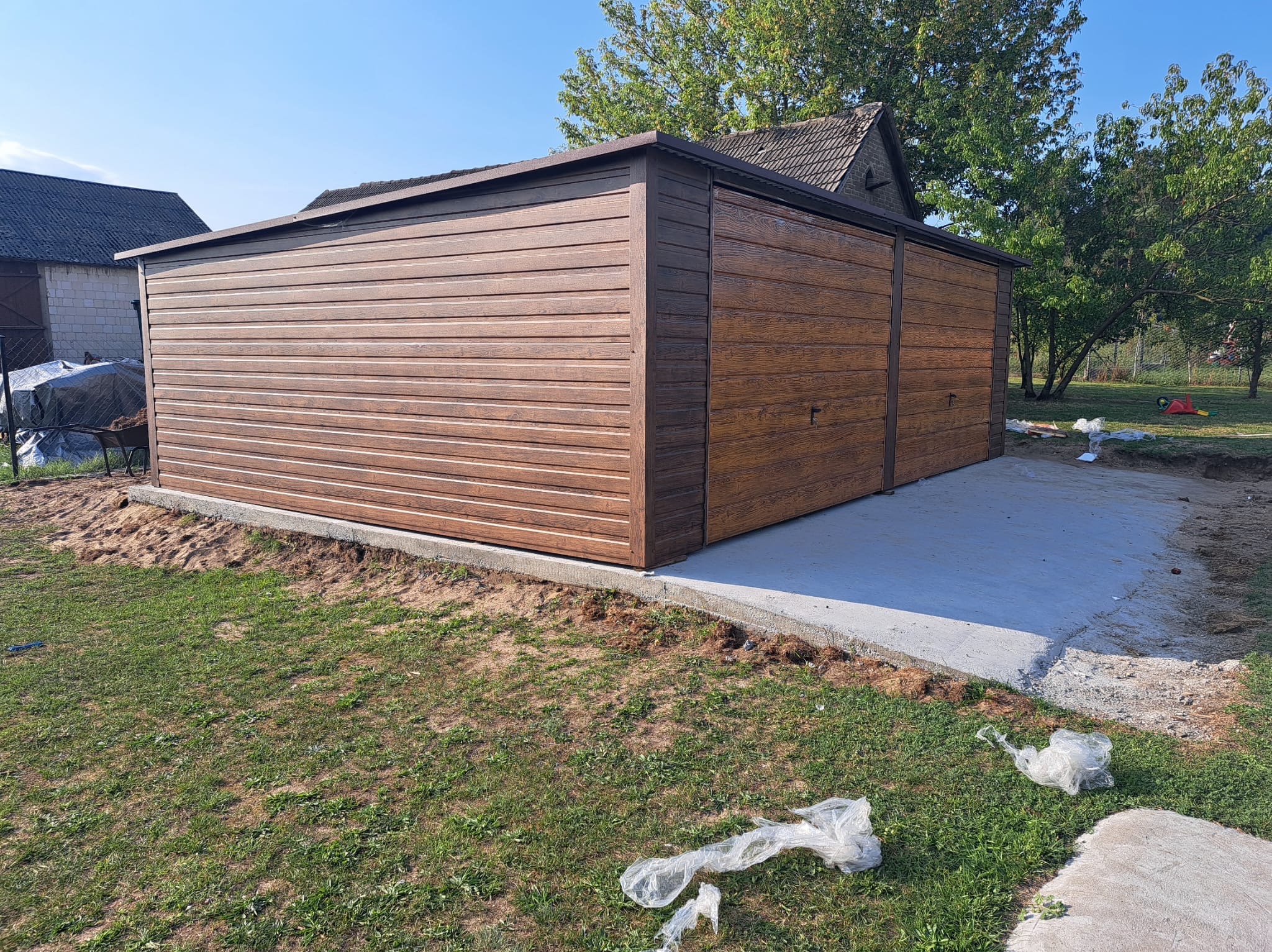 Dwustanowiskowy garaż blaszany z drewnopodobną elewacją, świeżo osadzony na betonowej wylewce, otoczony trawnikiem i zabudowaniami gospodarczymi.