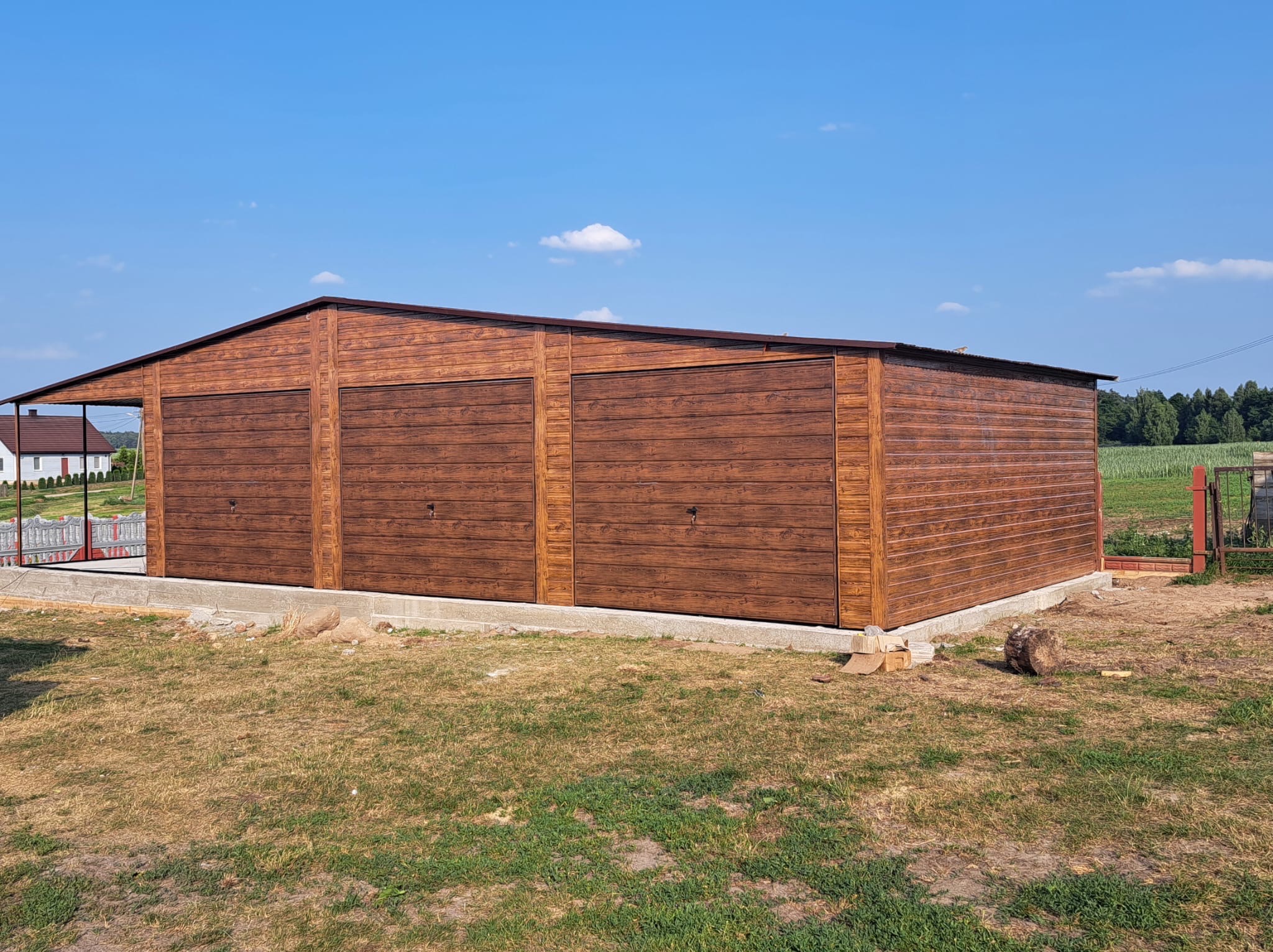 Trzysegmentowy garaż blaszany z bramami o teksturze drewna, osadzony na betonowej podstawie, na tle błękitnego nieba i zieleni.