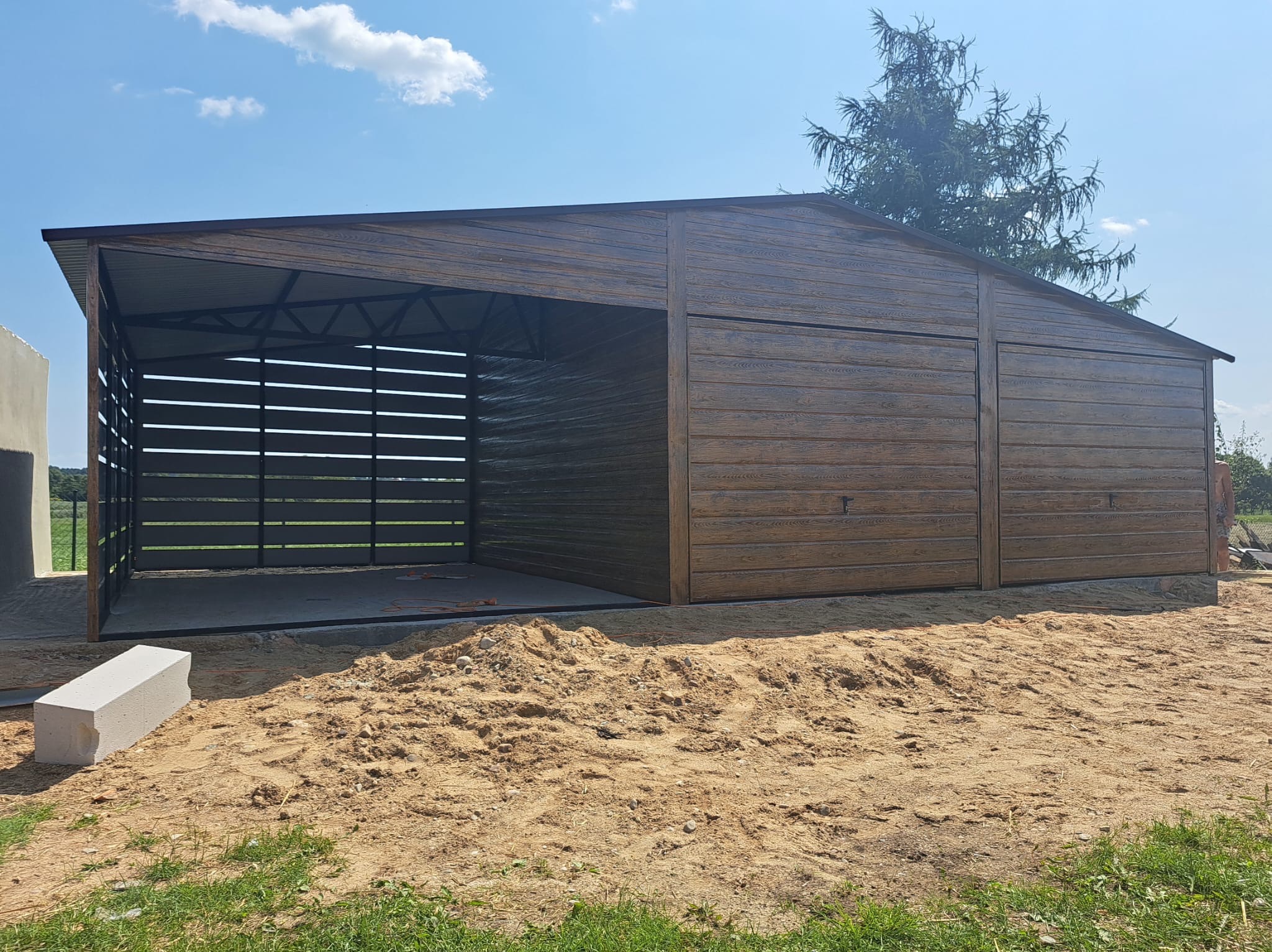 Drewnopodobny, otwarty garaż z metalową konstrukcją w Szczyrzycu. Widoczne elementy konstrukcyjne i boczna ściana z poziomych desek. Teren przygotowany pod wylewkę.