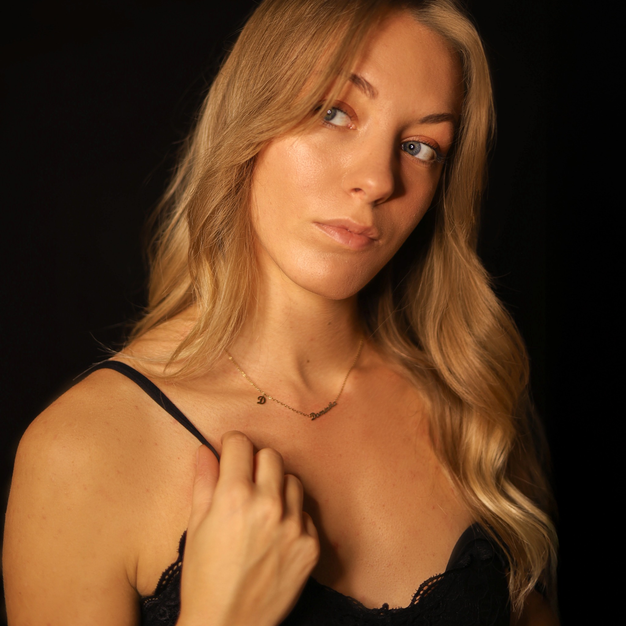 Portret młodej kobiety z blond włosami i niebieskimi oczami, delikatnie oświetlonej na ciemnym tle, z widocznym złotym naszyjnikiem z imieniem.