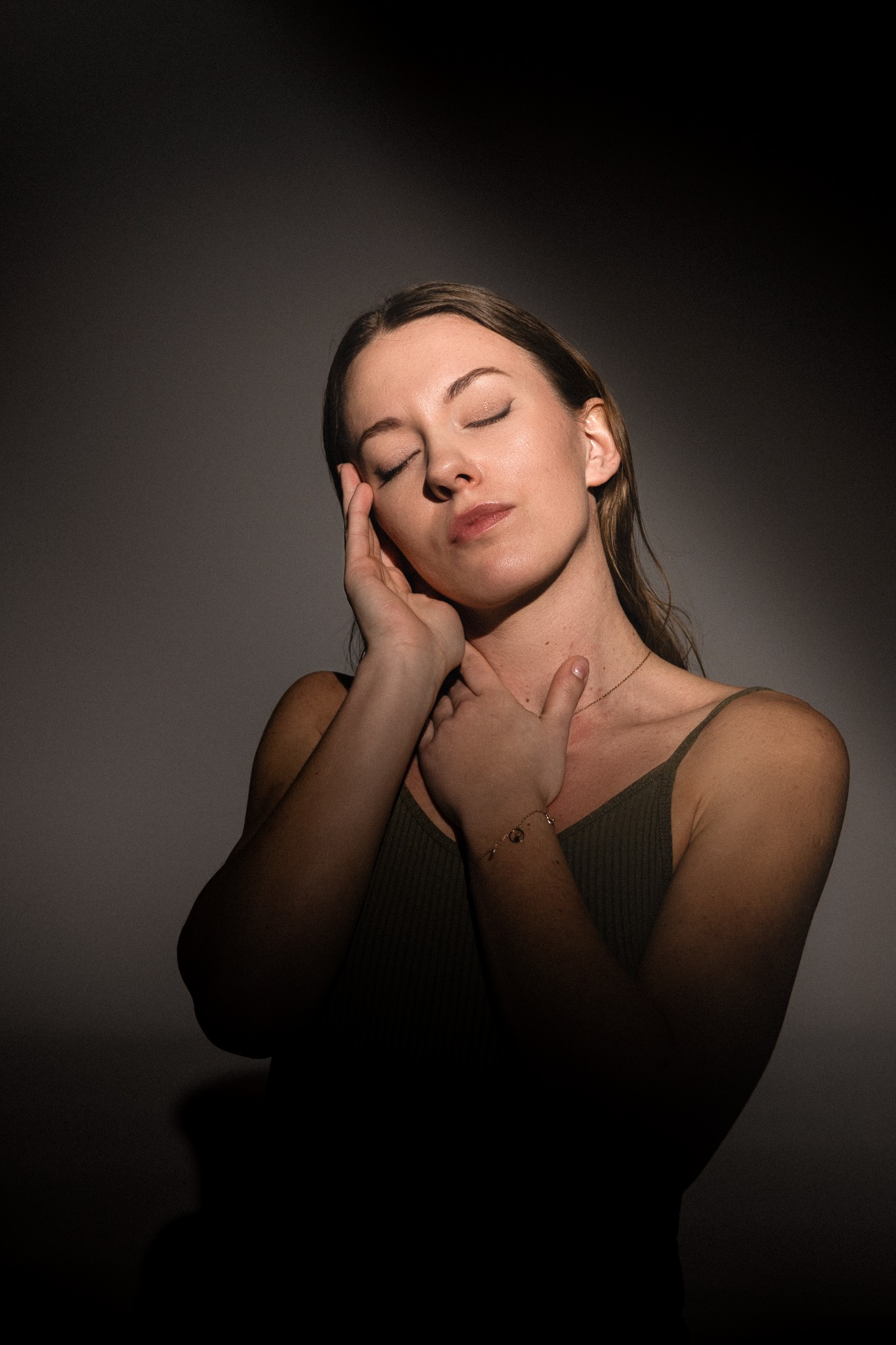 Portret kobiety z zamkniętymi oczami, delikatne oświetlenie podkreśla jej twarz i dekolt w zielonym topie, złoty łańcuszek i bransoletka dodają subtelności.